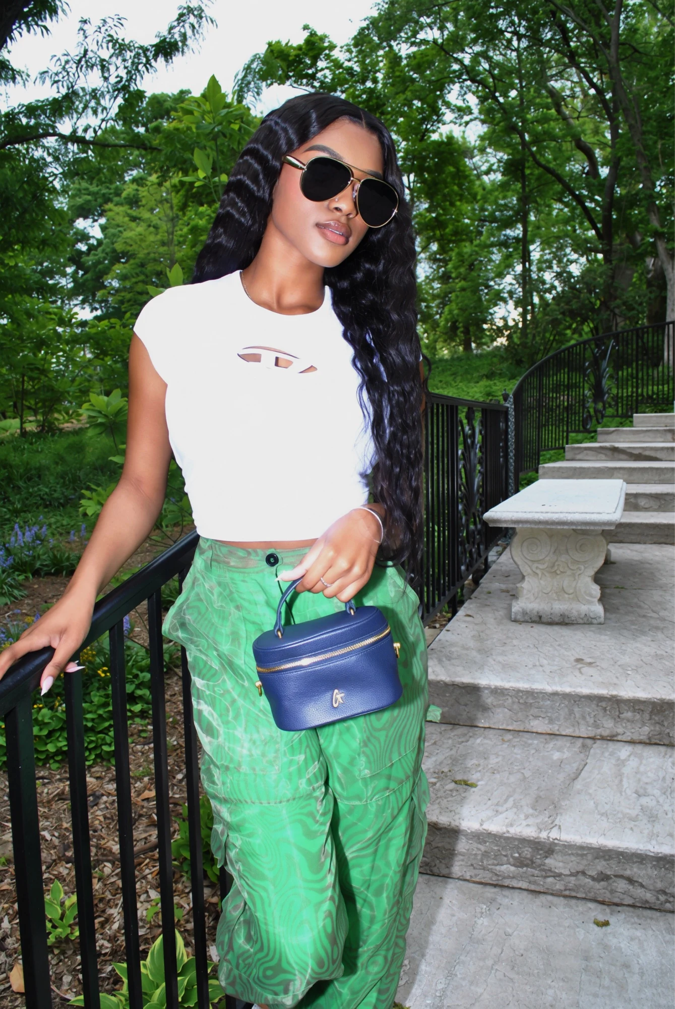 Sweet 16 garden portrait wearing sunglasses, a white top, and green patterned trousers, holding a blue handbag beside a stone staircase