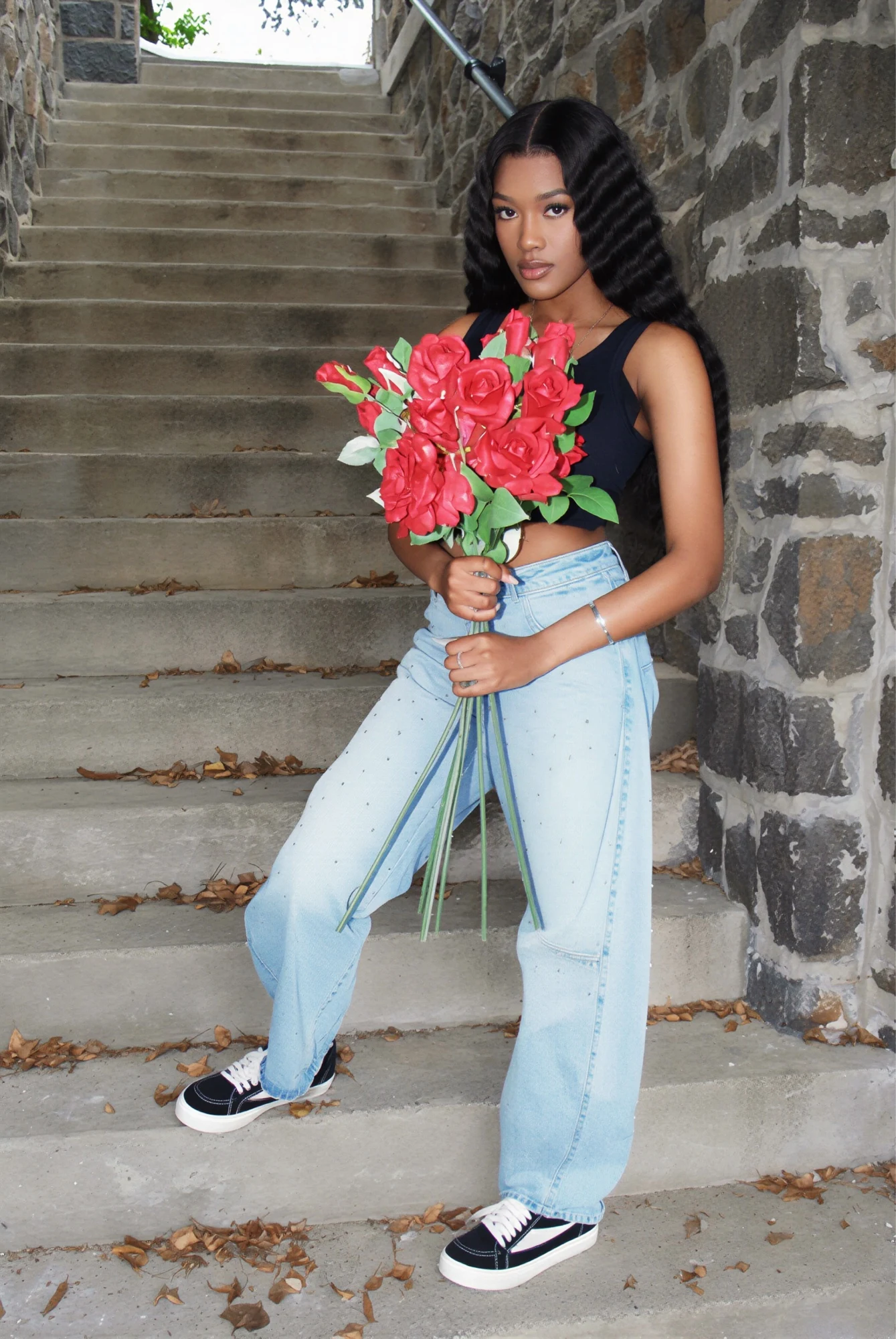 Sweet 16 portrait holding red roses on a stone staircase, wearing a black top, wide-leg jeans, and black sneakers